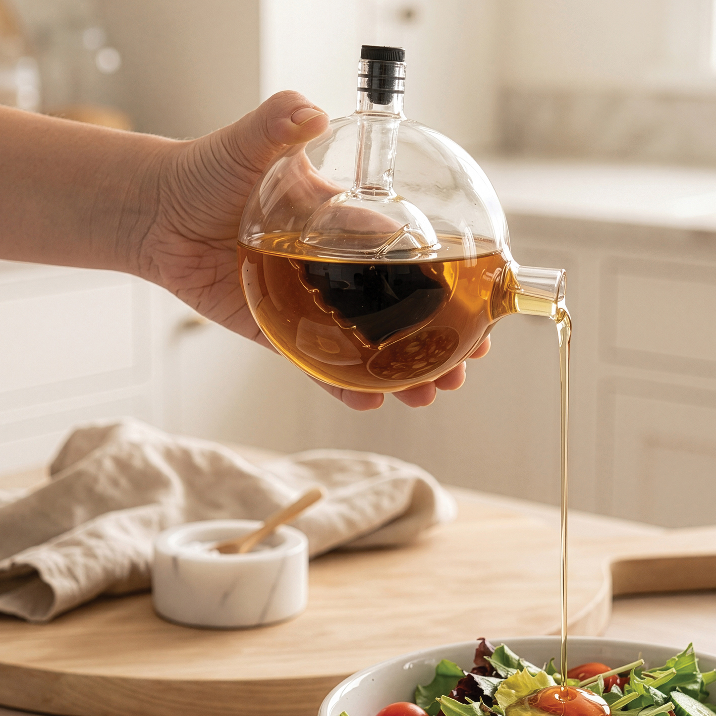 A hand pours oil from a Skully-Olio dispenser onto a salad in a bright kitchen.