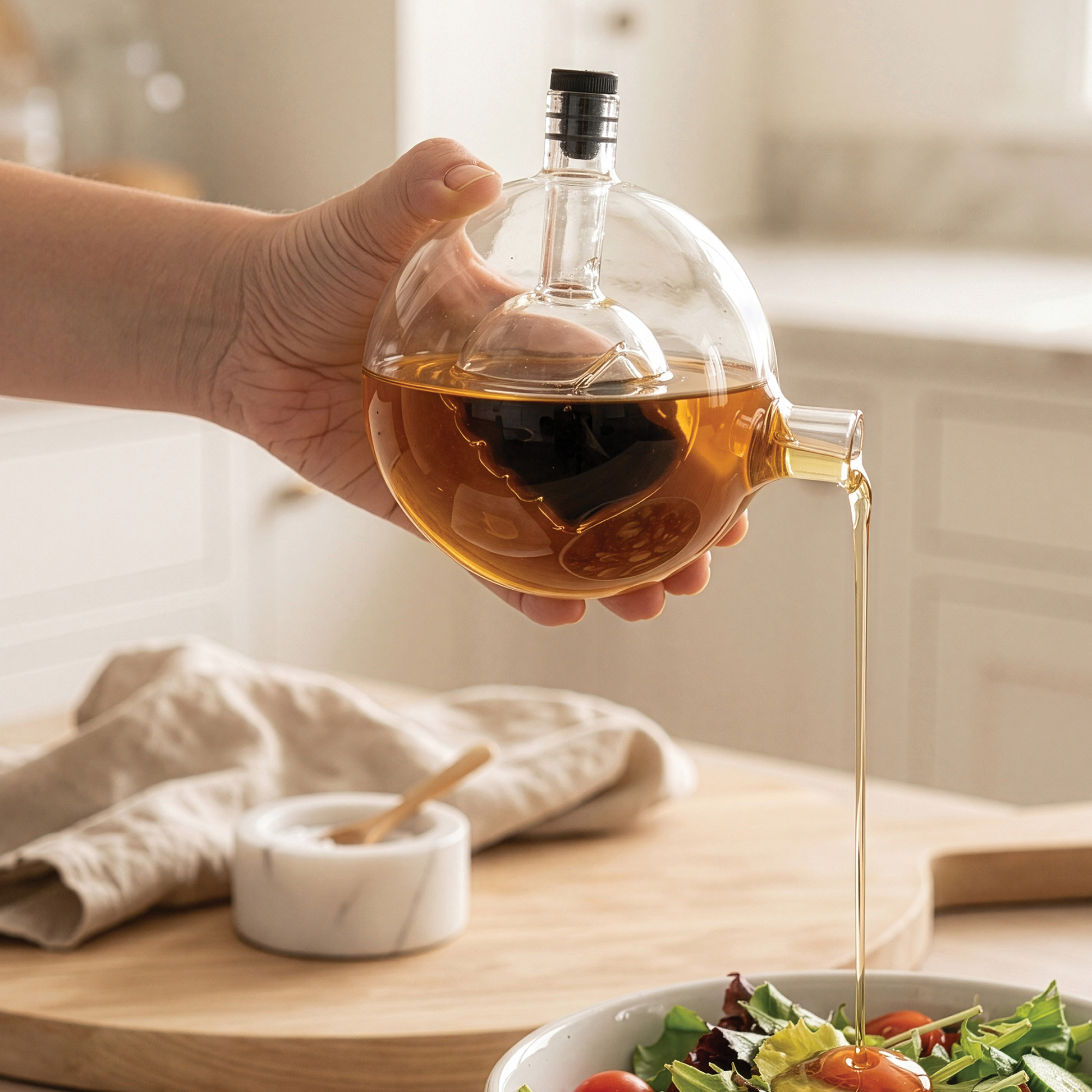 A hand pours oil from a Skully-Olio dispenser onto a salad in a bright kitchen.