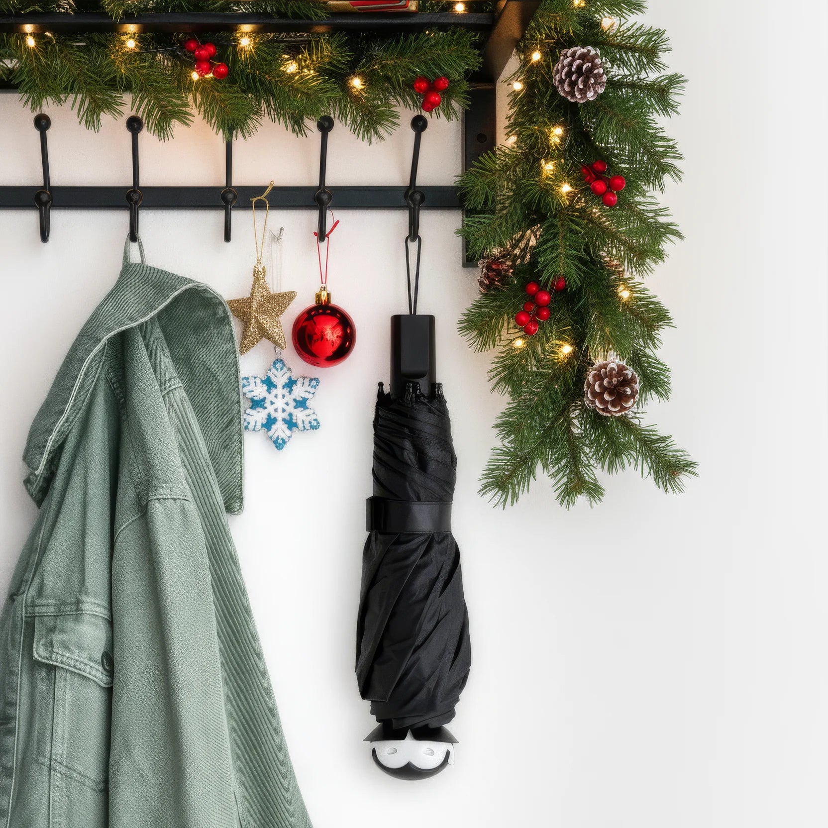 A green coat and a Spookula umbrella hang by a festive garland with red berries and ornaments.