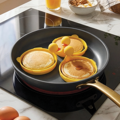 Three pancakes cooking in Sunny Side Duck egg rings on a stovetop; breakfast tools in background.