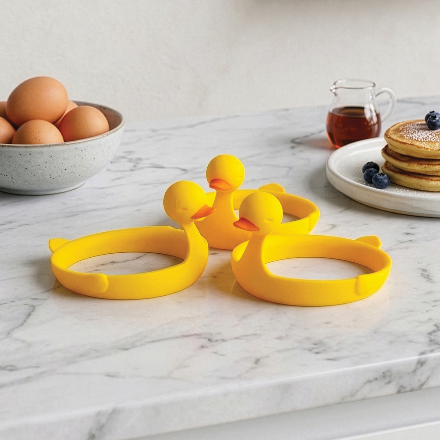 Three Sunny Side Duck egg rings sit on marble near eggs, pancakes, and syrup.