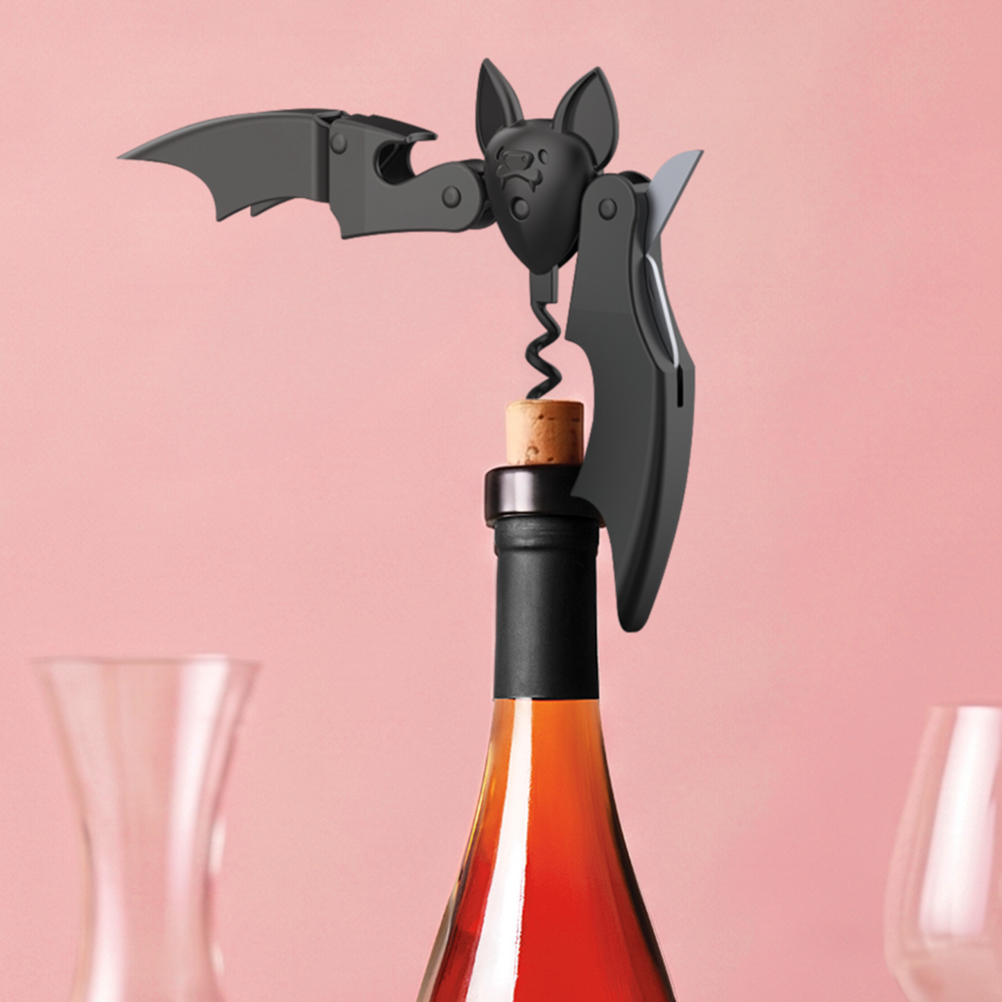 Vino bat-shaped OTOTO corkscrew opens a wine bottle, glassware softly blurred in the background.
