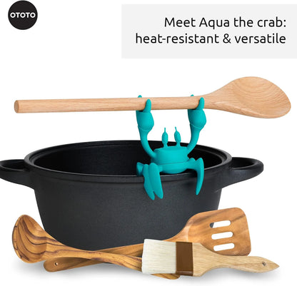 Aqua, the OTOTO crab utensil rest, grips a spoon on a pot with wooden kitchen tools nearby.