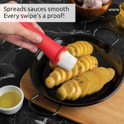 A hand uses a Gnomeo pastry brush to spread sauce on sliced potatoes in a black pan.