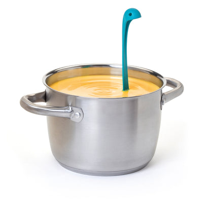 Nessie, a blue dinosaur-shaped OTOTO soup ladle, stands in a pot of yellow soup on white background.