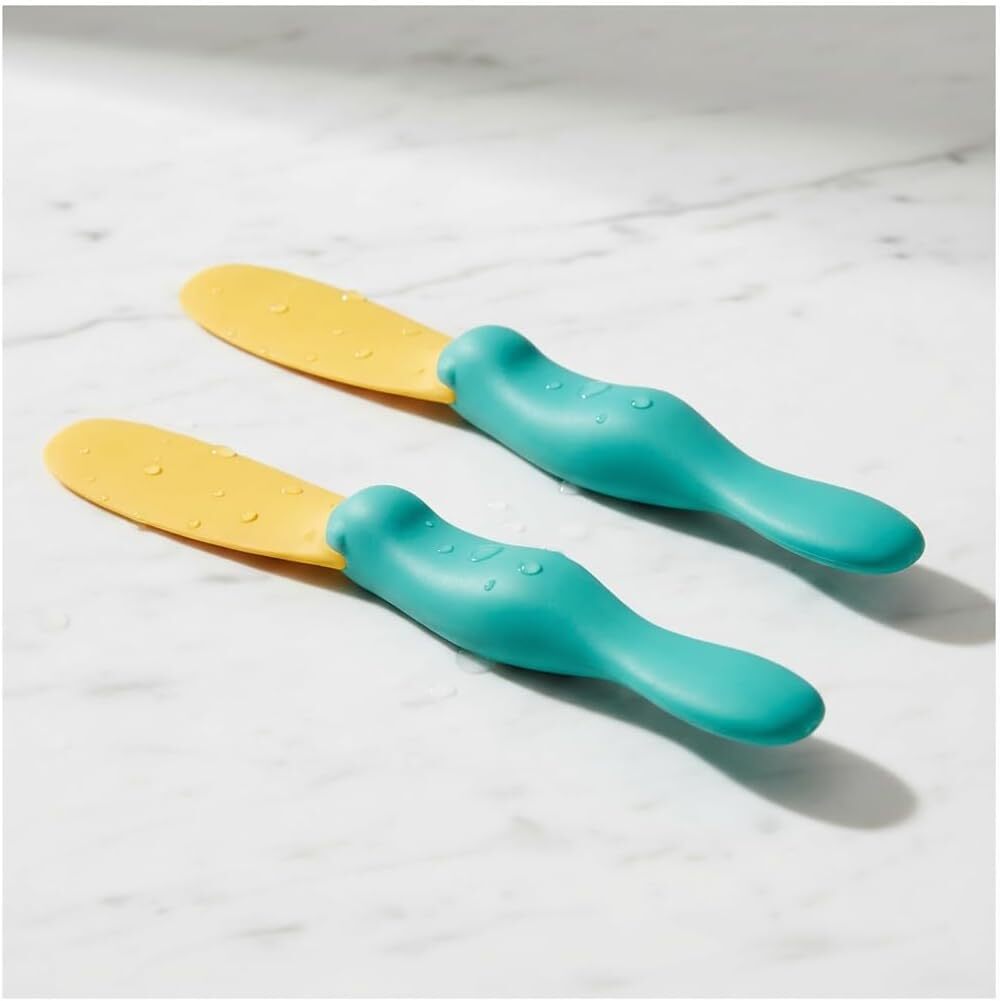 Two yellow and teal Splatypus spatulas with textured handles rest on a white marble surface.
