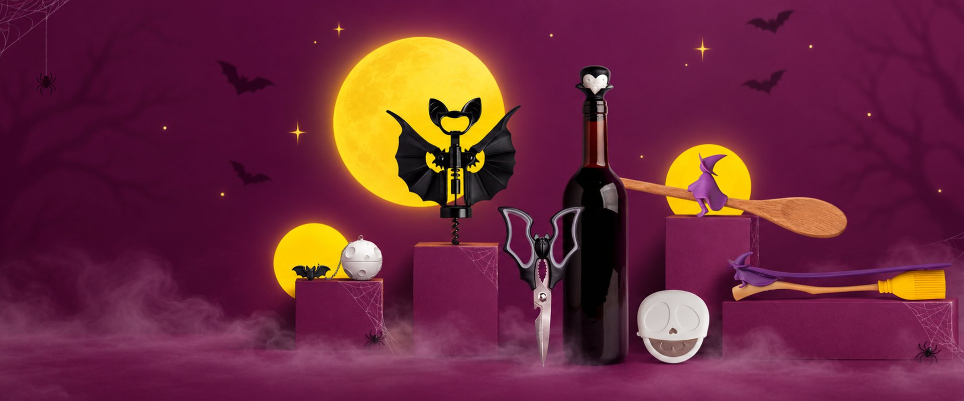 OTOTO's spooky kitchen on a purple set under a yellow moon: a bat corkscrew, bat scissors, a Dracula wine stopper, witch spoon rests and a skull