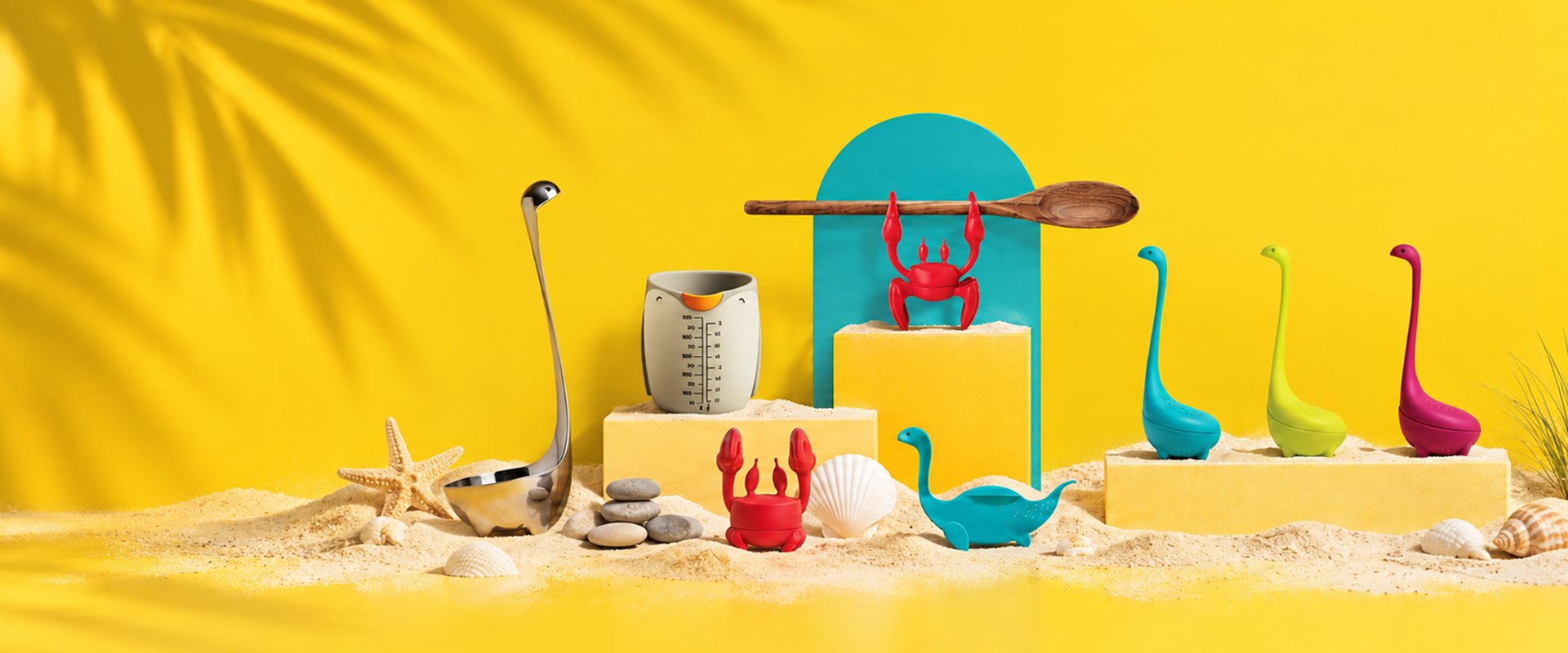 OTOTO best sellers on a sunny yellow beach set: Red the crab spoon holders, Nessie ladles and tea infusers, a steel Nessie ladle and a measuring cup