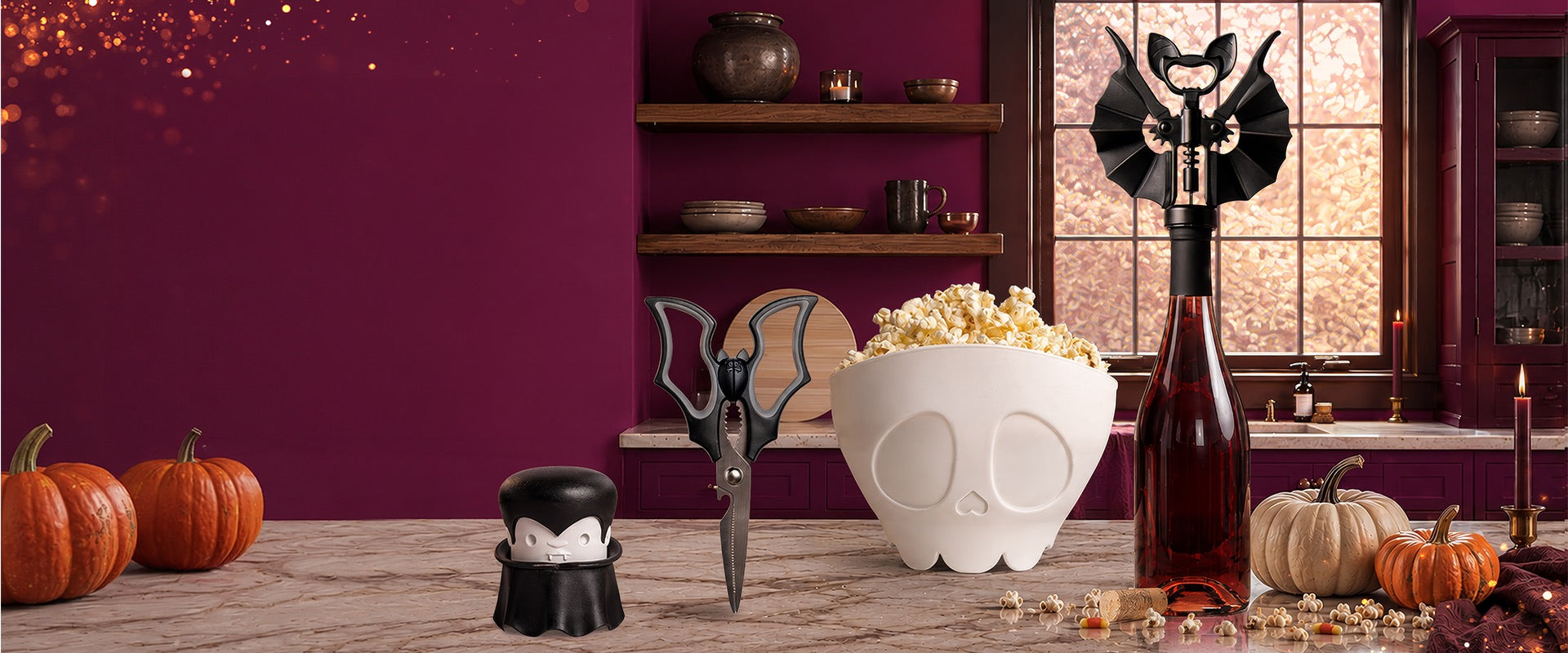 OTOTO's spooky kitchen on a marble counter with pumpkins: a Dracula garlic crusher, bat kitchen scissors, a skull popcorn bowl and a bat corkscrew on a bottle of red wine