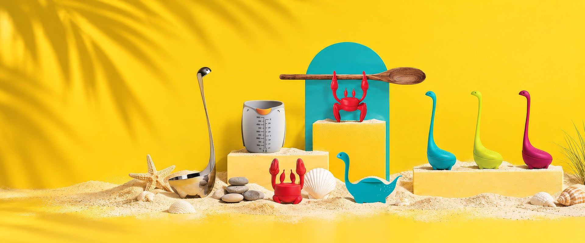 OTOTO best sellers on a sunny yellow beach set: Red the crab spoon holders, Nessie ladles and tea infusers, a steel Nessie ladle and a measuring cup
